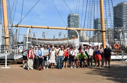 Group of tourists posing on a harbor promenade in front of a tall-sailed ship with masts and rigging, city skyline in the background.