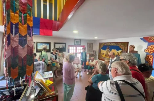 Group of elderly people in a cultural room listening to a guide beside colorful wall hangings and display cases.