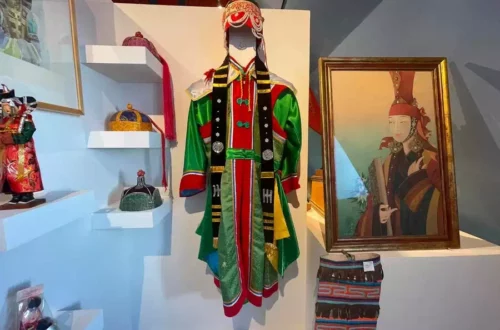 Colorful traditional costume on a mannequin in a museum display, with a headdress and black vest, flanked by framed art and woven textiles.