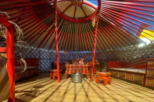 Interior of a red-and-gold yurt with central hanging stove, carved wooden tables and benches, and lattice walls.