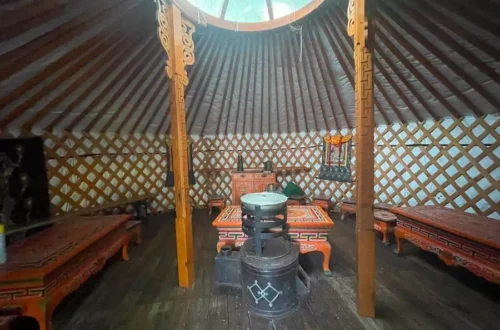 Interior of a traditional yurt with lattice walls, carved wooden pillars, and a central stove surrounded by low orange-patterned benches and tables under a round skylight.