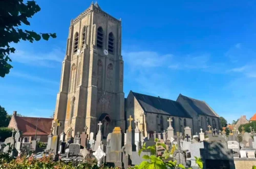 Stone church with a tall square bell tower beside a graveyard under a blue sky; crosses and headstones in the foreground.