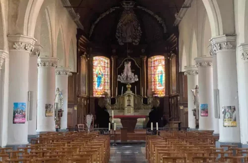 Interior of a church nave with wooden pews facing an ornate altar and stained-glass windows at the front.