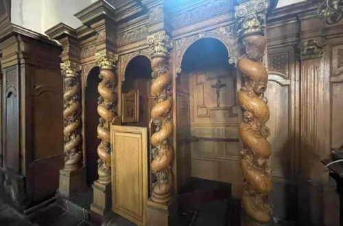 Wooden pulpit with elaborately twisted carved columns in a church interior, cross visible on the wall behind.