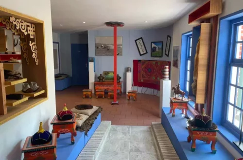 A bright museum room with traditional artifacts on low pedestals, colorful textiles, and framed photos on light blue walls. A red central pole stands in the middle.