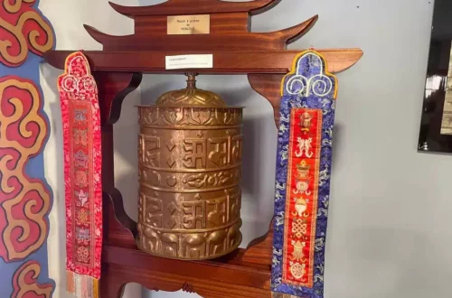 Bronze Tibetan prayer wheel mounted in a carved wooden frame with red and blue fabric banners on either side, decorative and ceremonial display.