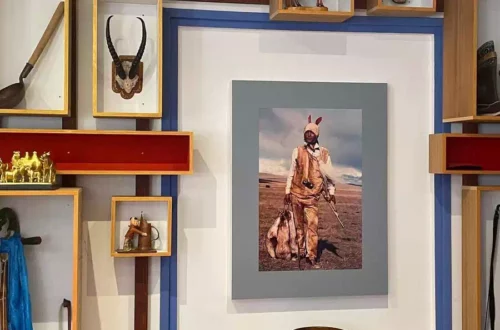 Framed photo of a hunter in tan gear with a rabbit-ear headpiece, holding a rifle beside a game bag in a dry, open field.